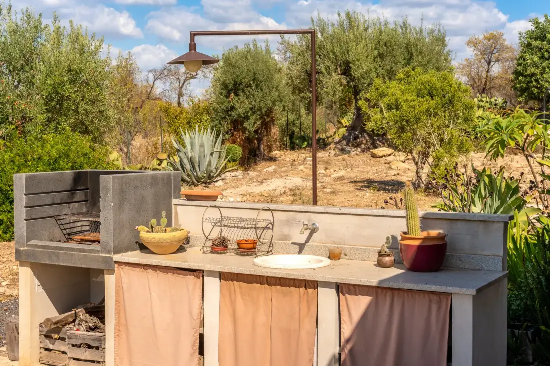 Una encimera de cocina al aire libre con fregadero y parrilla está rodeada de arbustos y árboles.