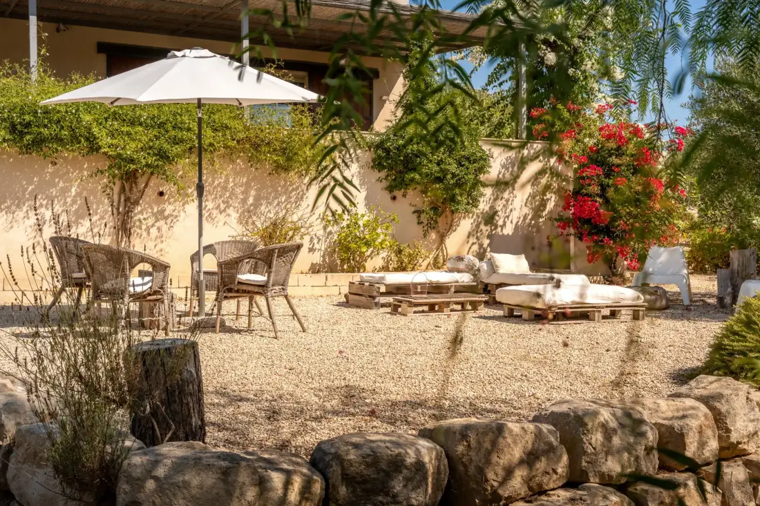 Un patio ajardinado de grava con asientos al aire libre, una sombrilla blanca y plantas con flores.