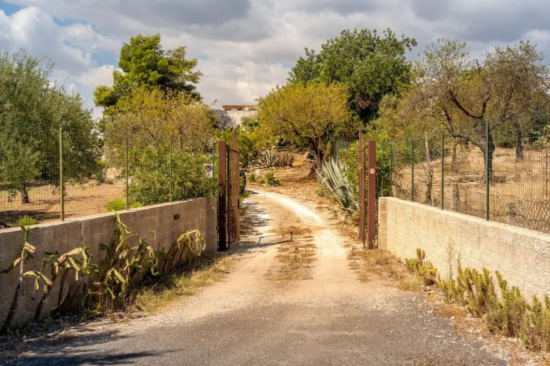 Un camino de tierra pasa por una puerta abierta entre muros bajos y árboles.