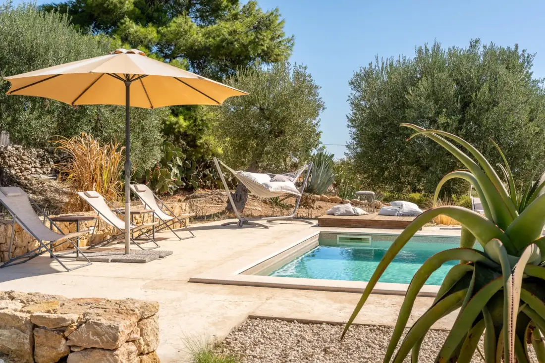 Una piscina al aire libre junto a un patio soleado con asientos y una sombrilla.