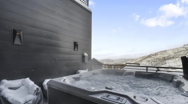 An outdoor hot tub sits beside a dark building, with snowy hills in the distance.