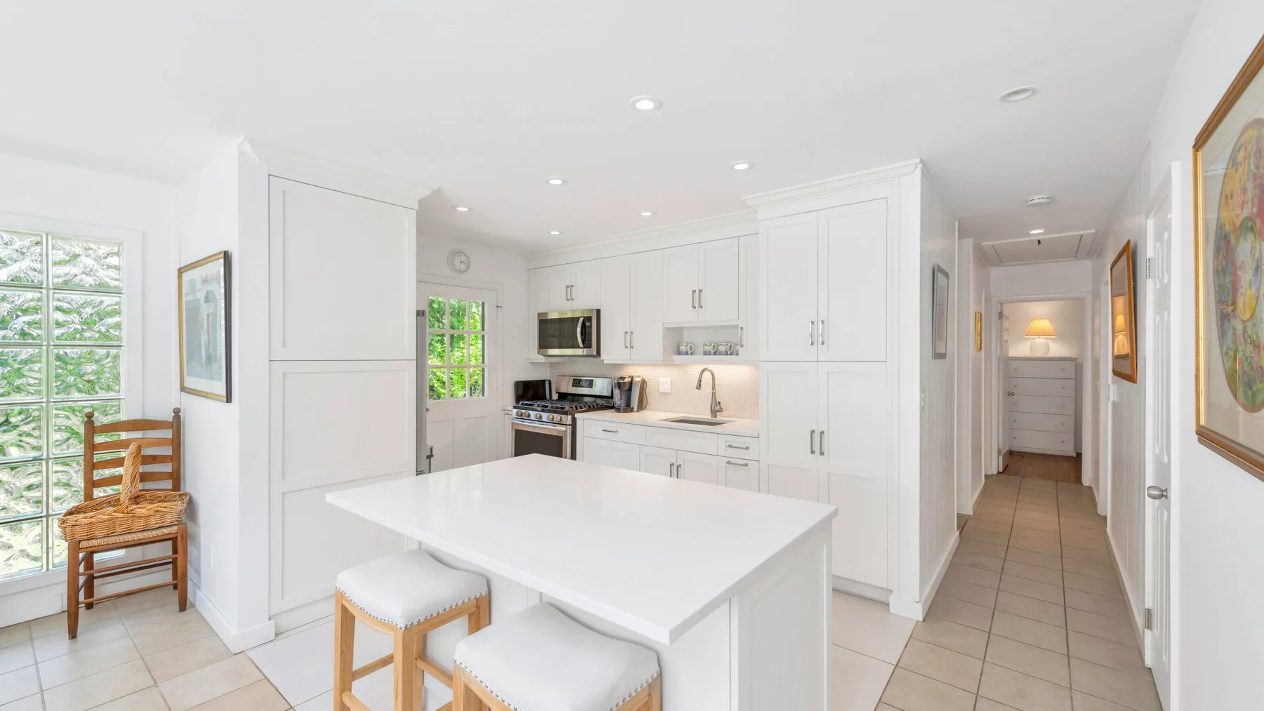 Una cocina luminosa con gabinetes blancos, una isla central y un pasillo contiguo.