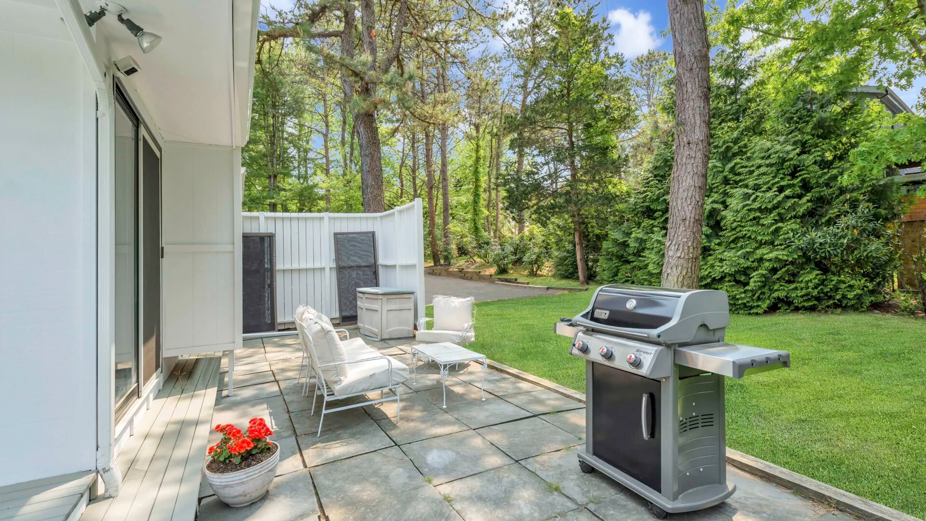 Un patio exterior pavimentado junto a una casa tiene una parrilla y asientos frente a un jardín verde.