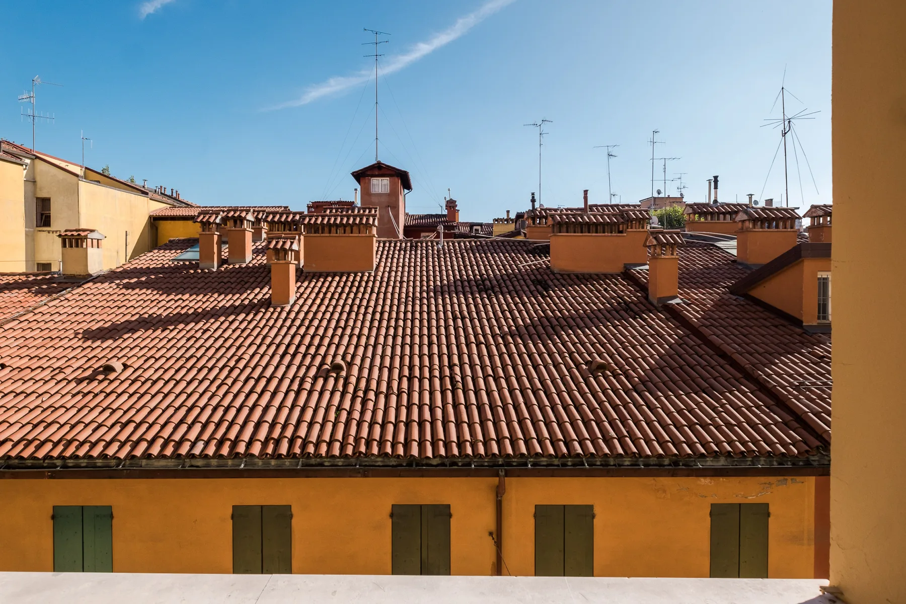 Rooftop view of terracotta tiles, chimneys, and antennas beneath a clear sky.