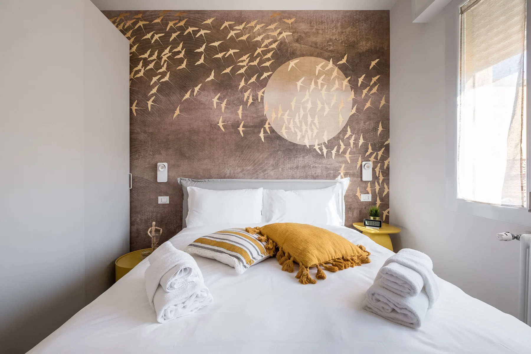 A bright bedroom has a neatly made bed beneath a mural of flying birds.