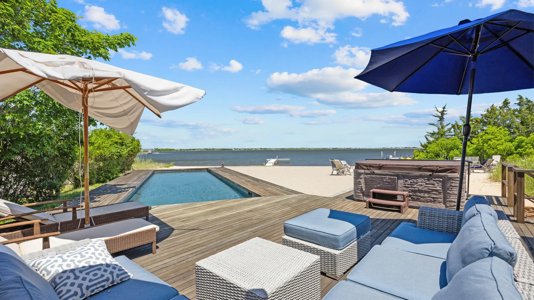 A waterfront patio has lounge seating, umbrellas, and a pool overlooking the water.