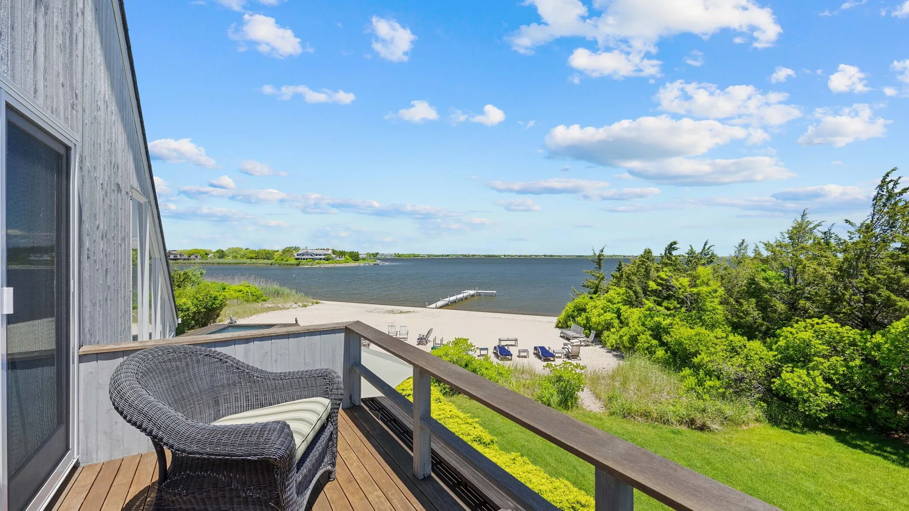 A deck overlooks a sandy beach and calm water, with outdoor seating nearby.
