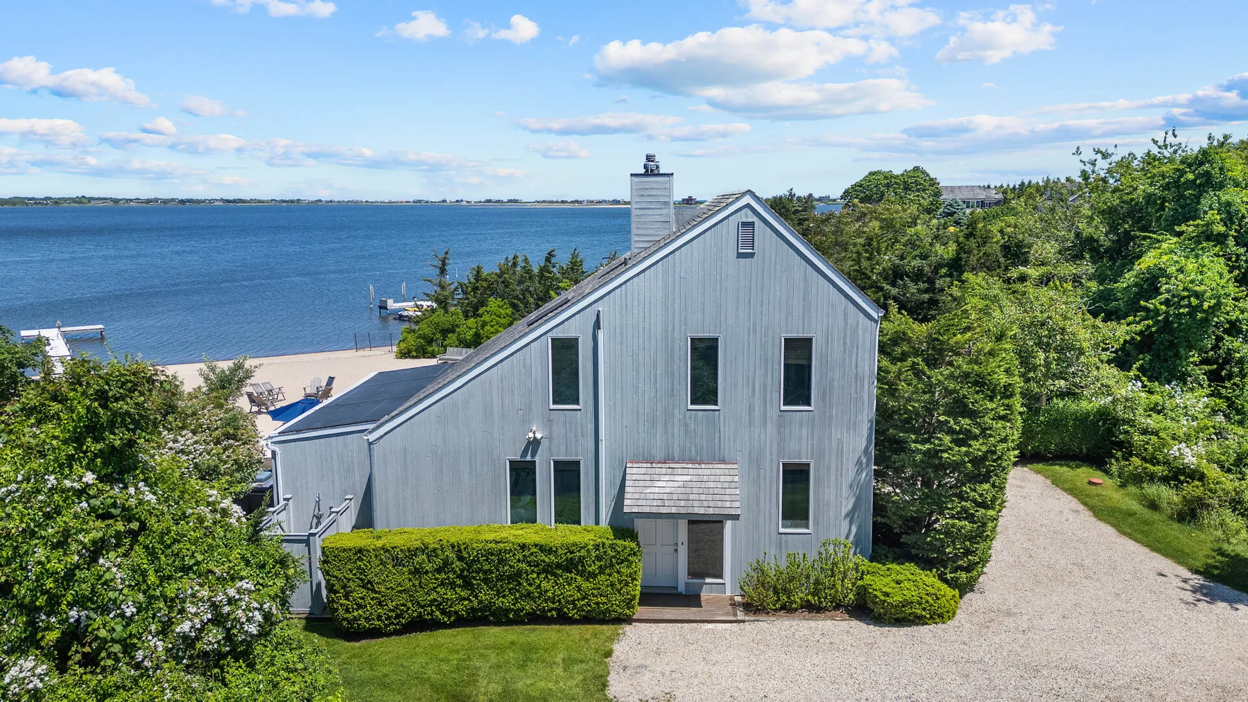 A gray house beside a body of water, with trees and a gravel drive.