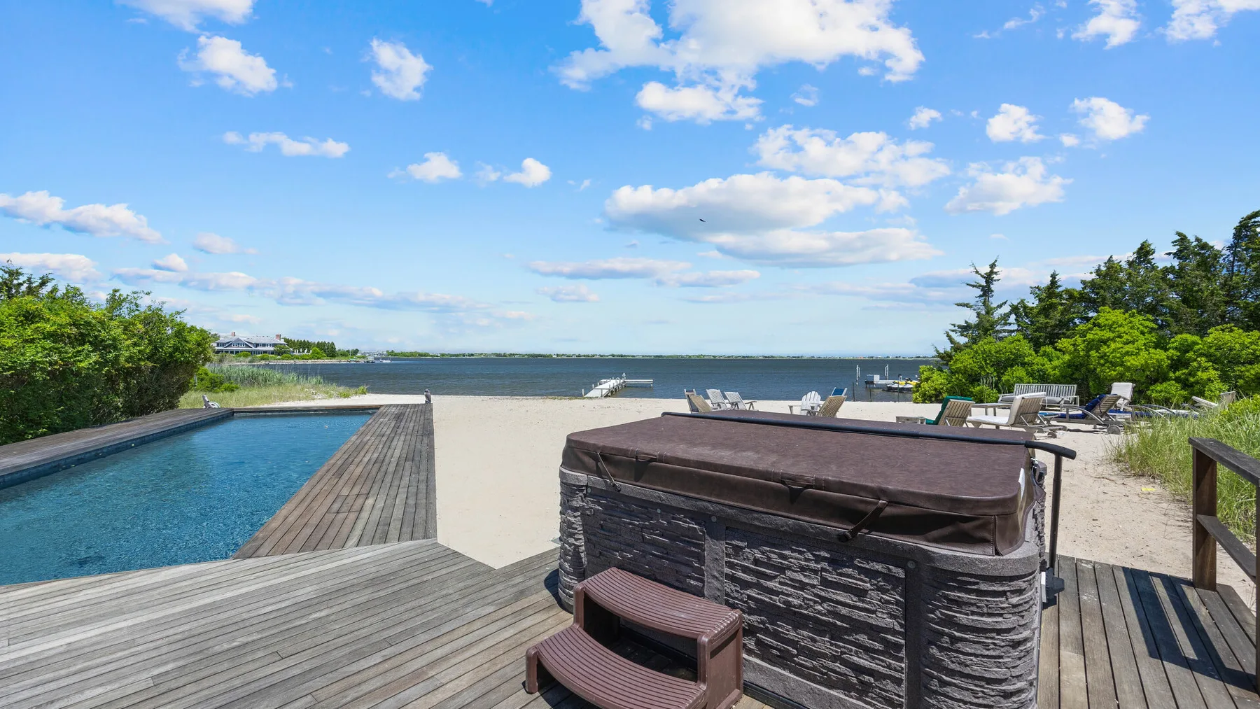 An outdoor hot tub sits on a wooden deck beside a swimming pool overlooking the water.