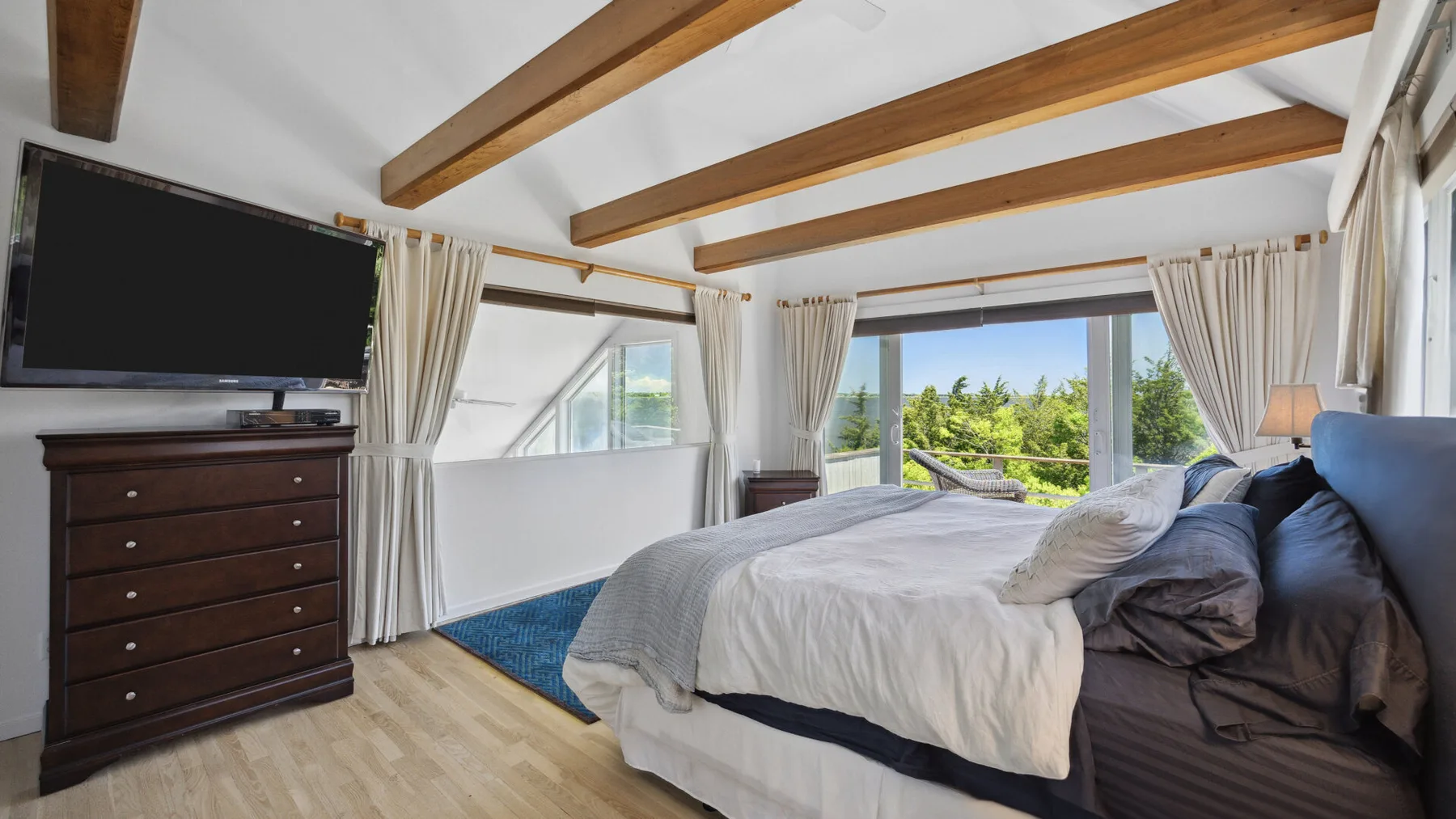 A bedroom with a bed, a dresser, and large windows overlooking greenery.