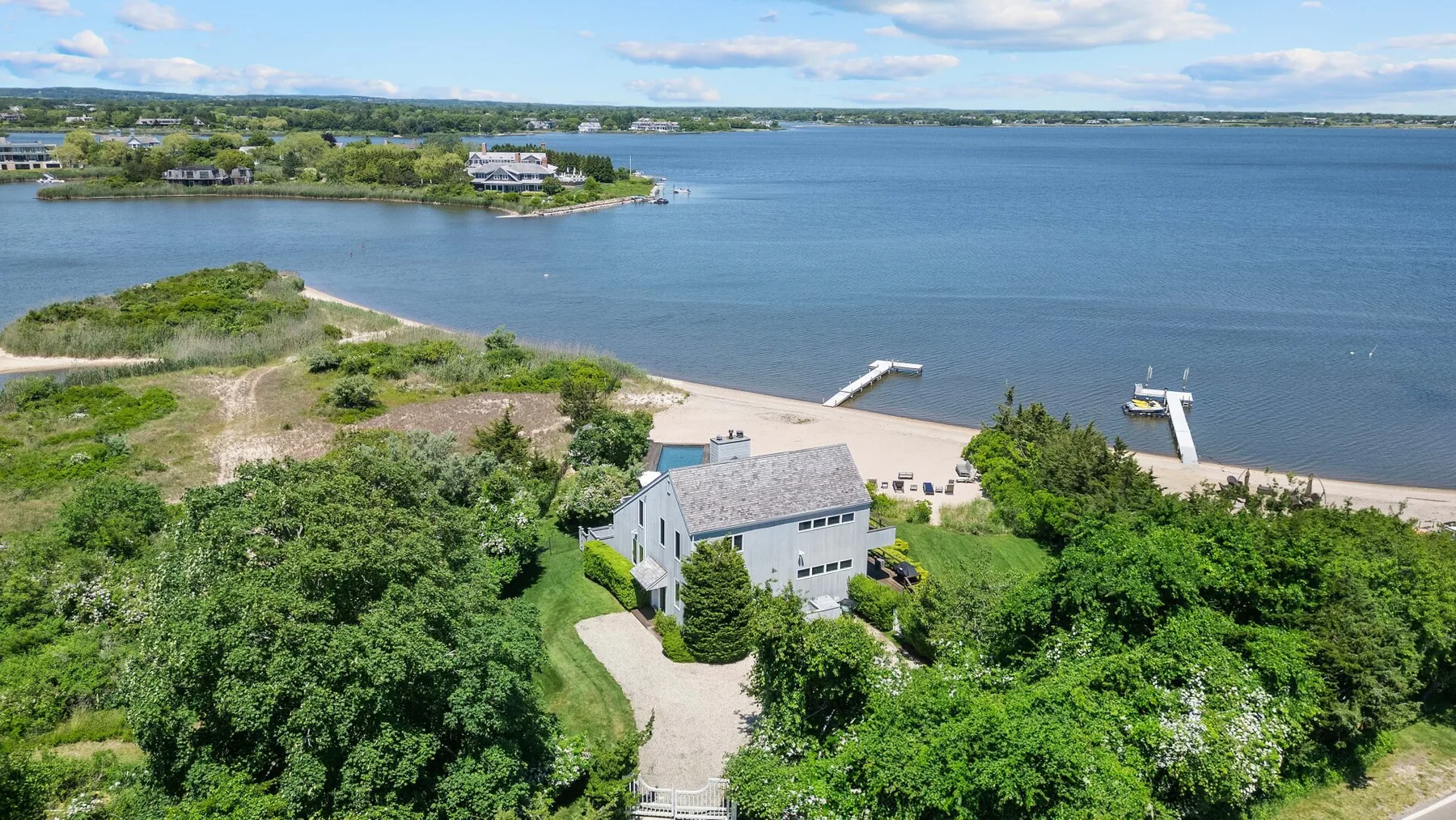A white house sits among green trees beside a broad lake and sandy shore.