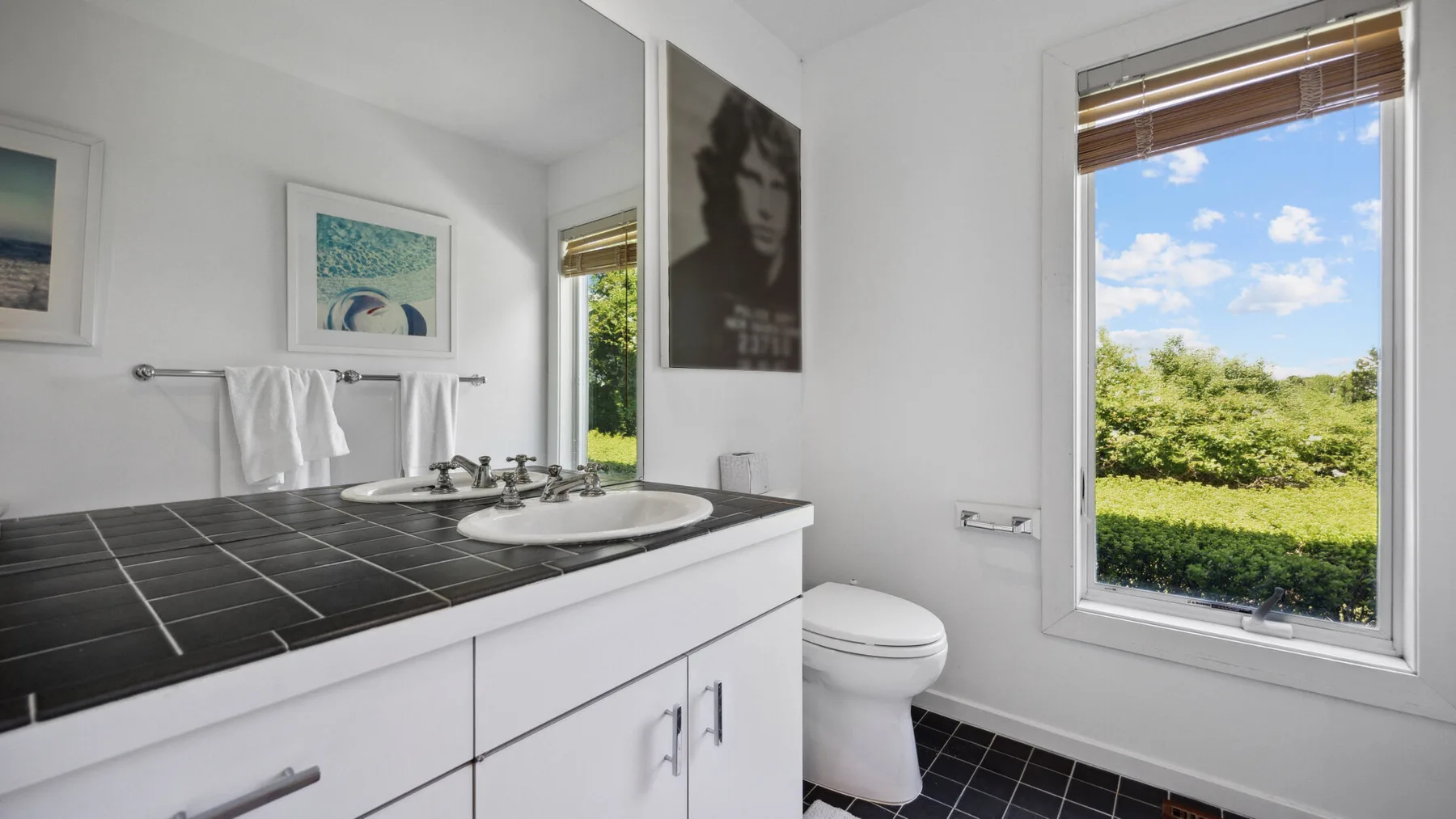 A bright bathroom with a dark tiled vanity and a large window overlooking greenery.
