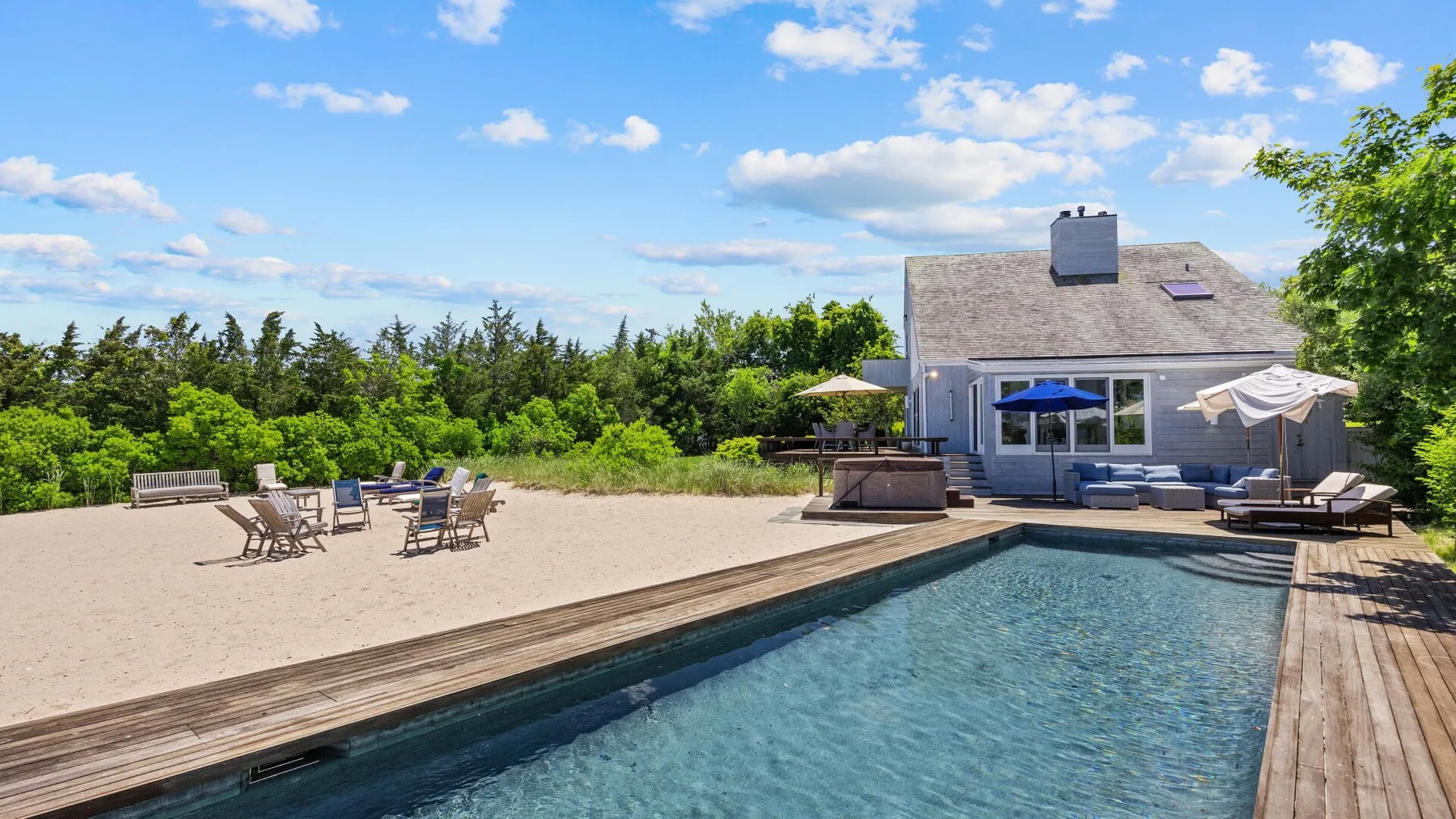 A swimming pool and sandy patio sit beside a house, surrounded by trees.