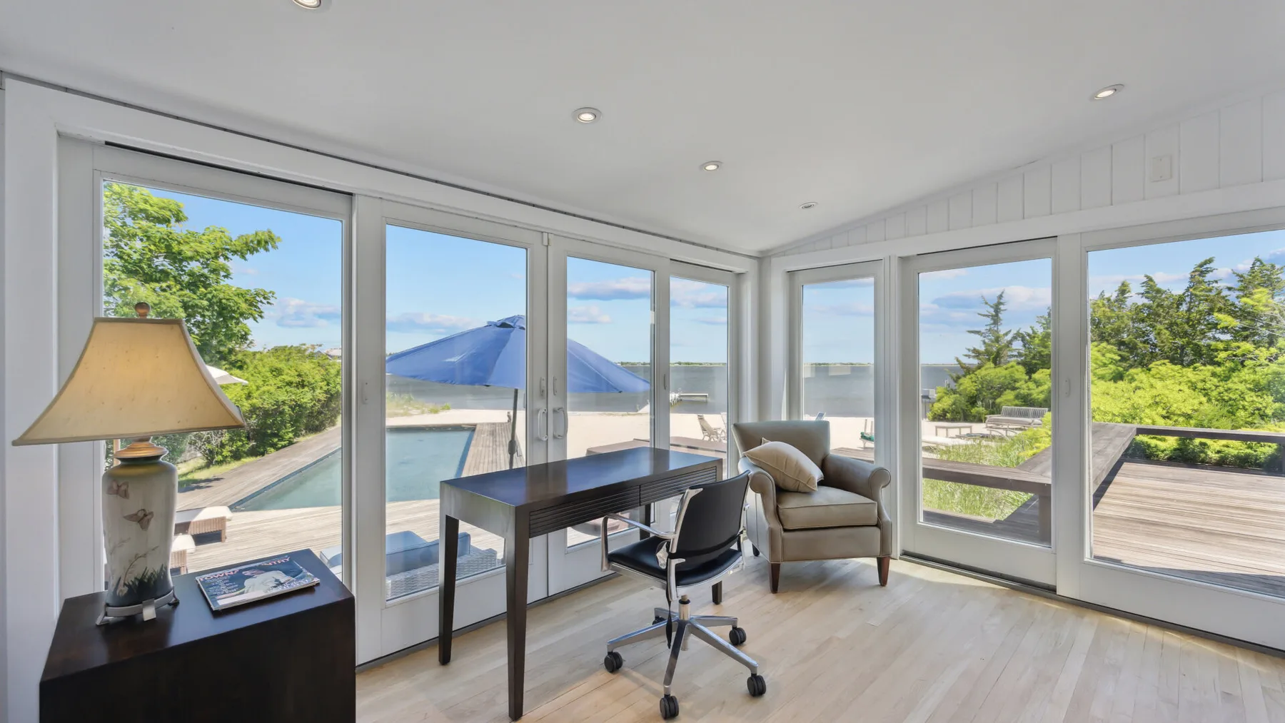 A bright room with a desk and seating beside large windows overlooking a pool and greenery.