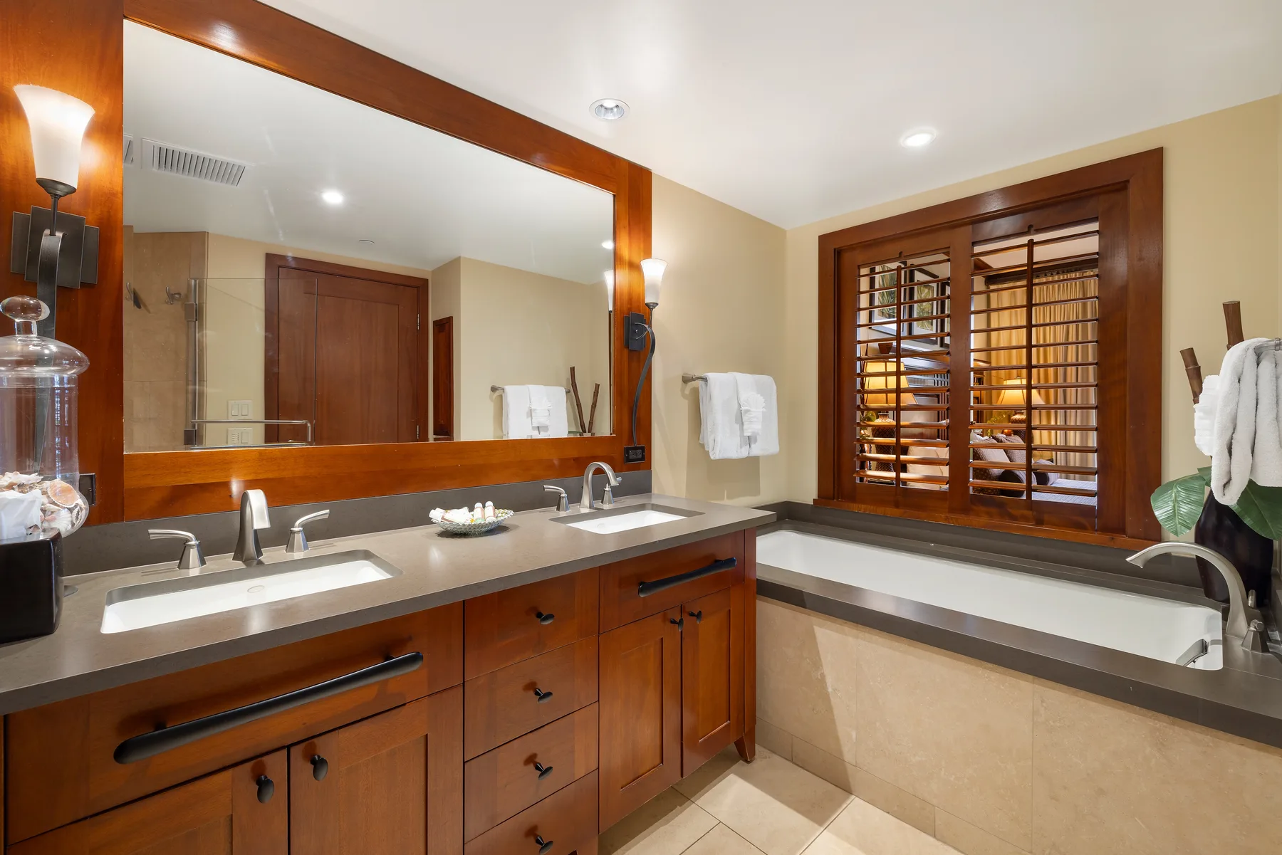 Un tocador de baño con un espejo amplio y una bañera bajo ventanas con persianas.