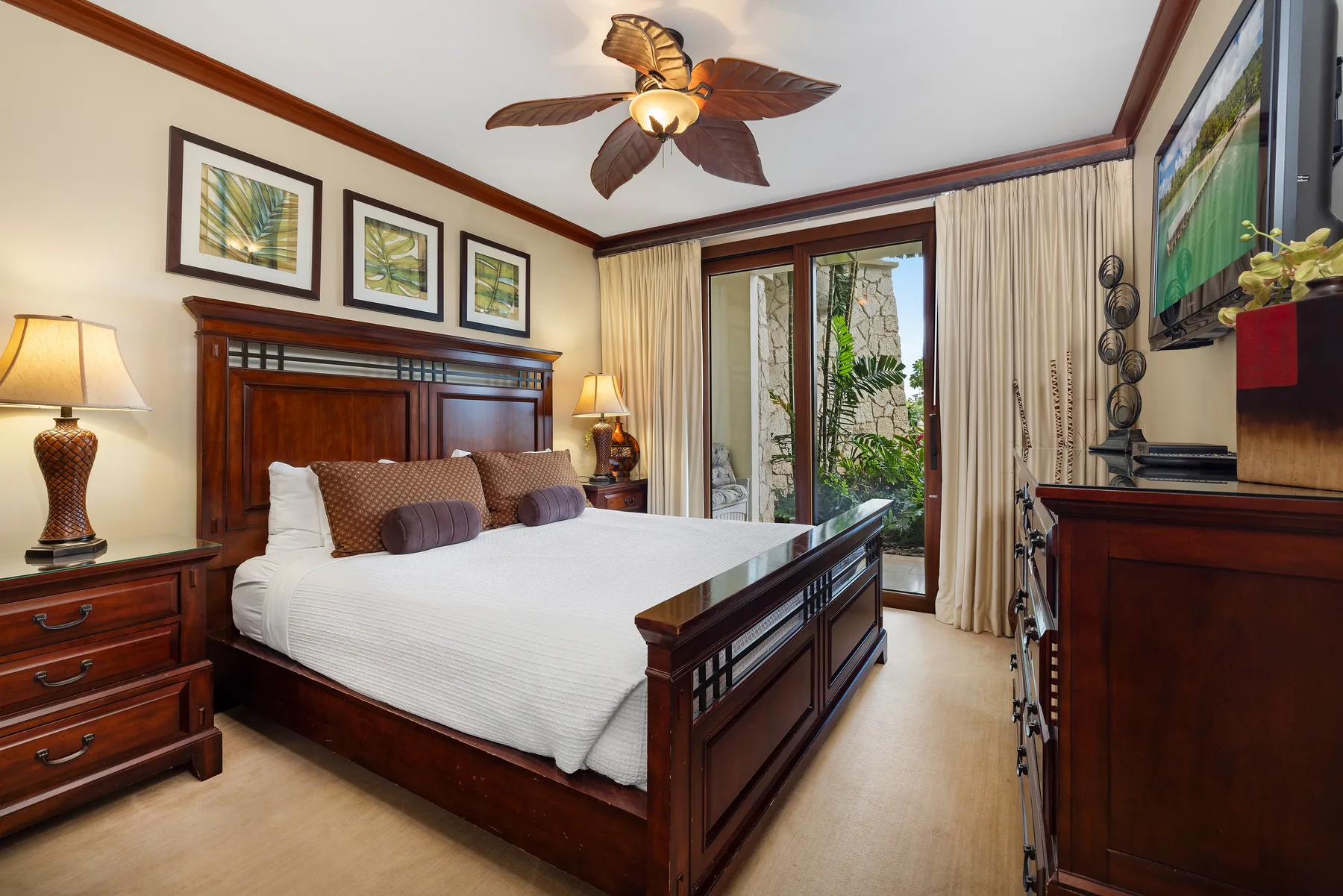 Un dormitorio con una cama grande de madera, cuadros enmarcados y puertas de vidrio hacia un jardín.