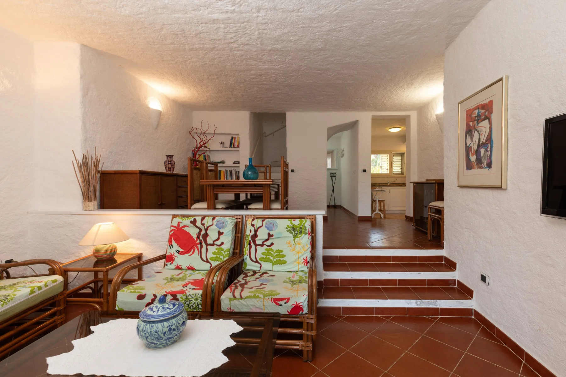 A living area with patterned seating and a dining table beyond a short tiled stair.