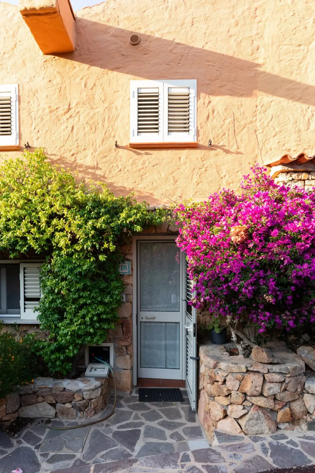 A stucco house entrance opens onto a stone-paved path framed by flowering shrubs.