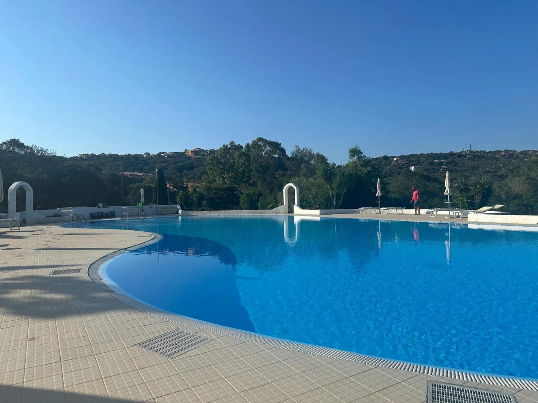 A large outdoor swimming pool is surrounded by a tiled deck and wooded hills.