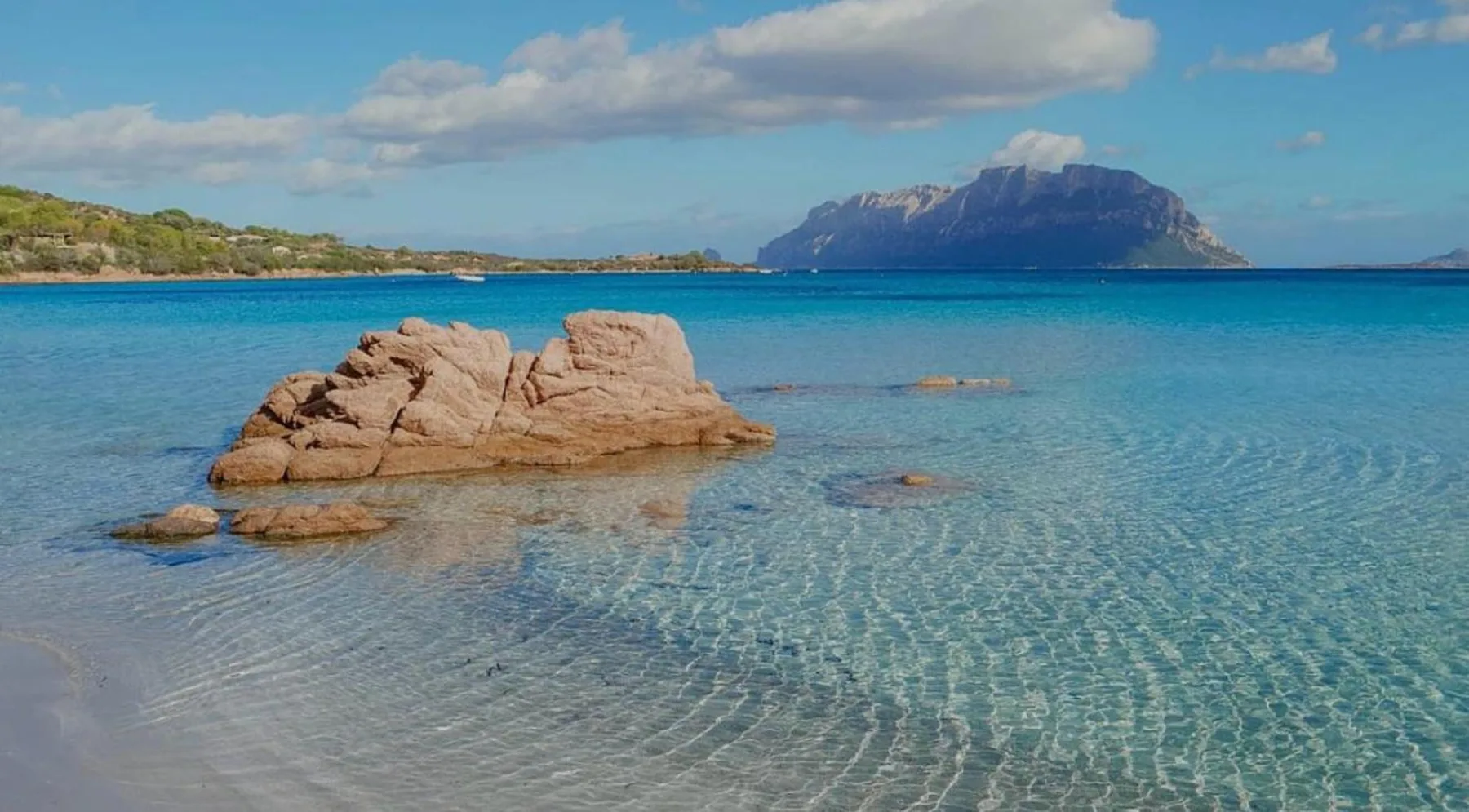 A clear blue bay with rocky outcrops and a mountainous island in the distance.