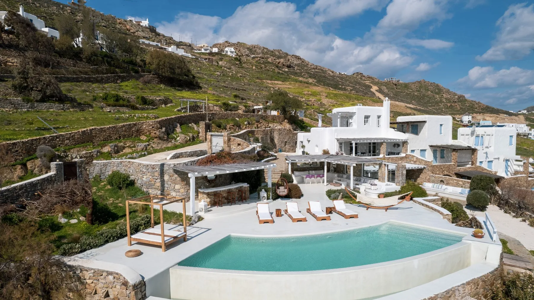 A white hillside villa overlooks a curved pool and outdoor seating.
