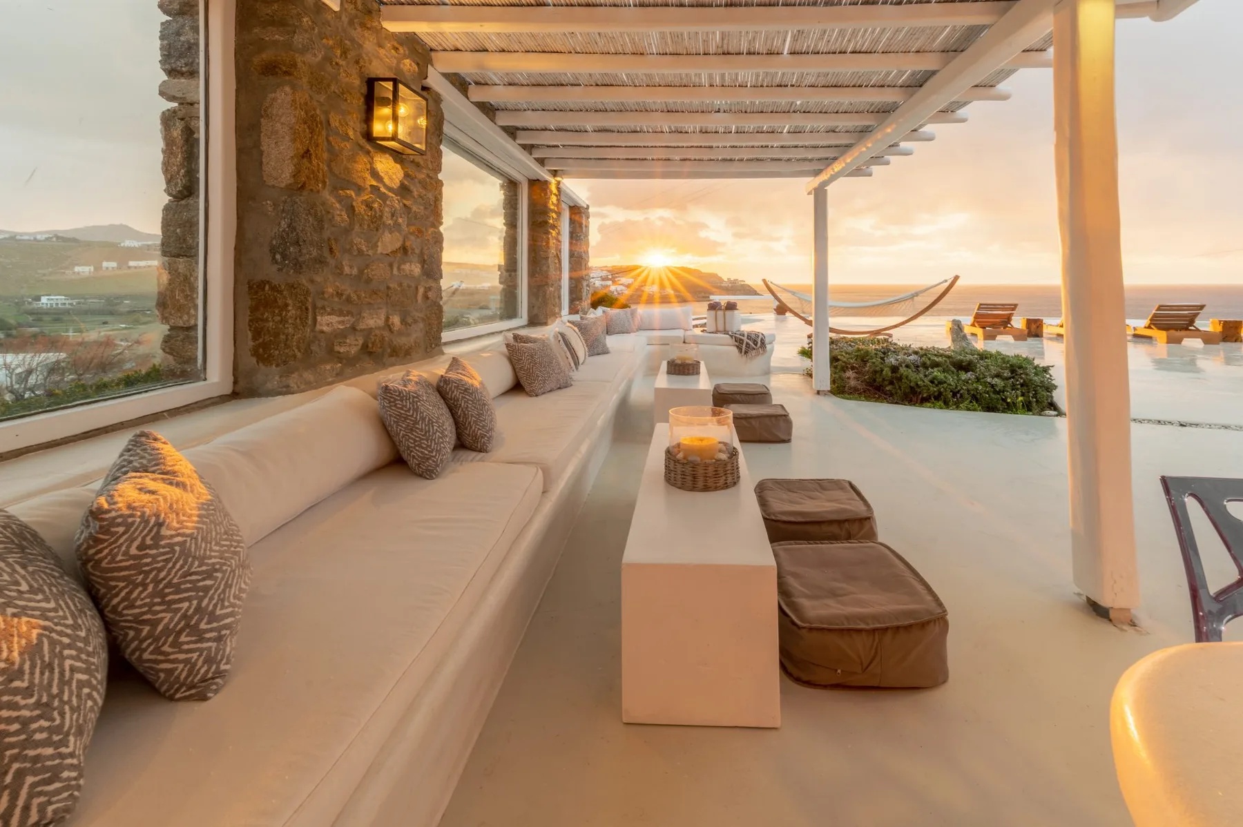 A covered outdoor lounge with cushioned seating overlooks the water at sunset.