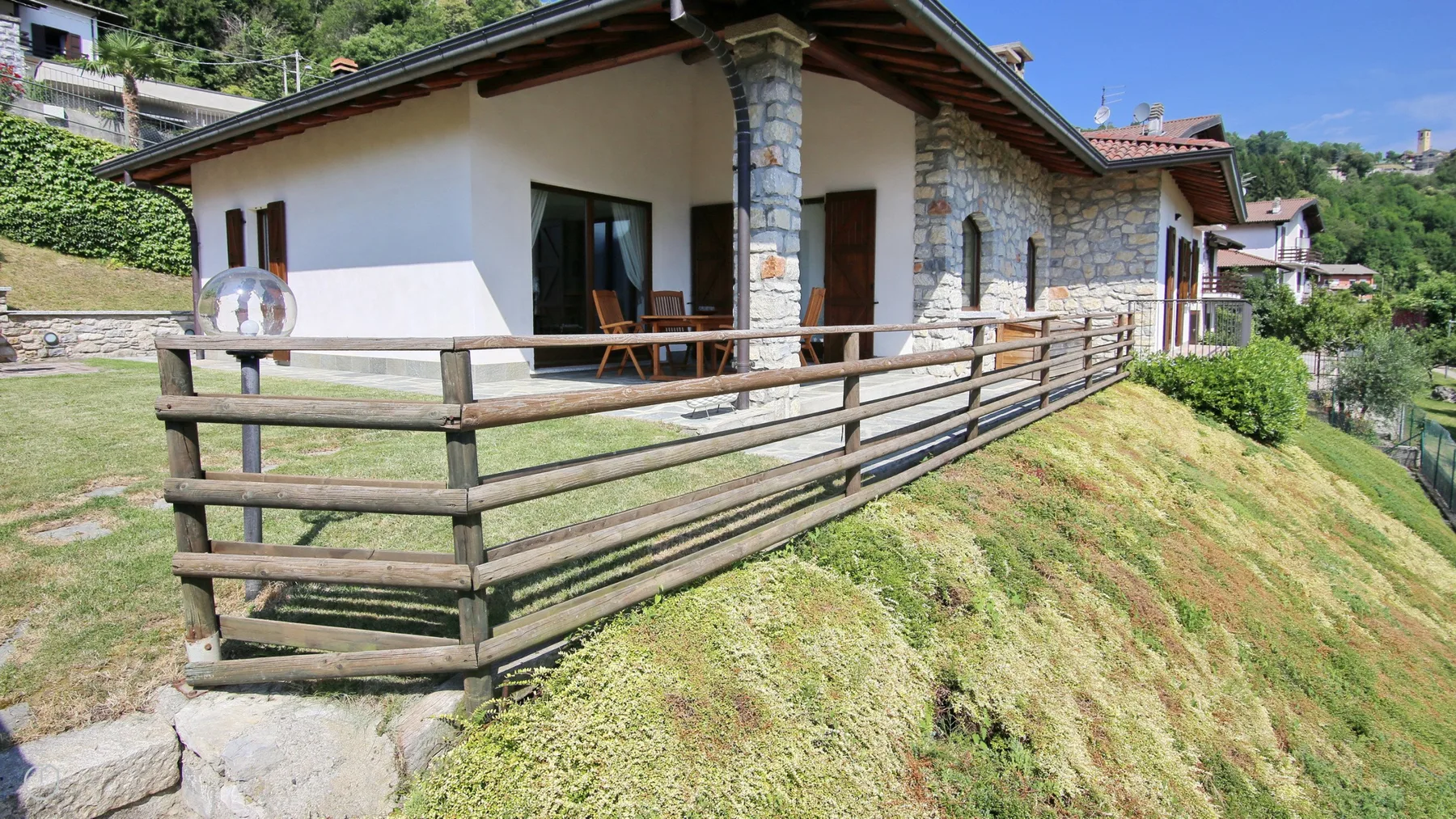 A stone-and-stucco house has a long wooden-railed terrace on a grassy slope.