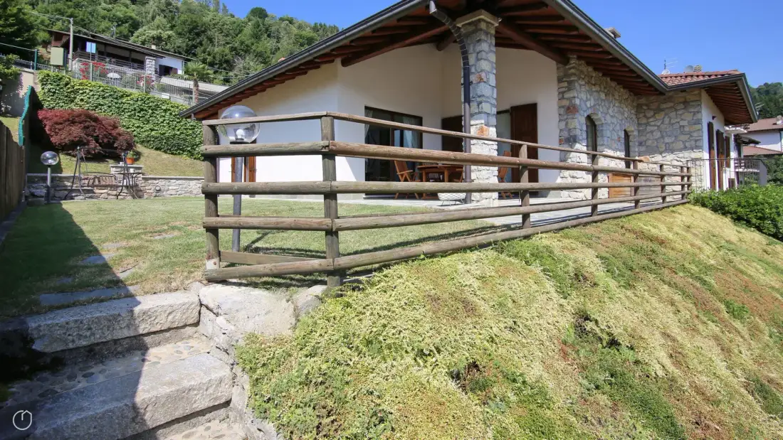 A stone-and-plaster house with a wooden railing beside the sloping lawn.