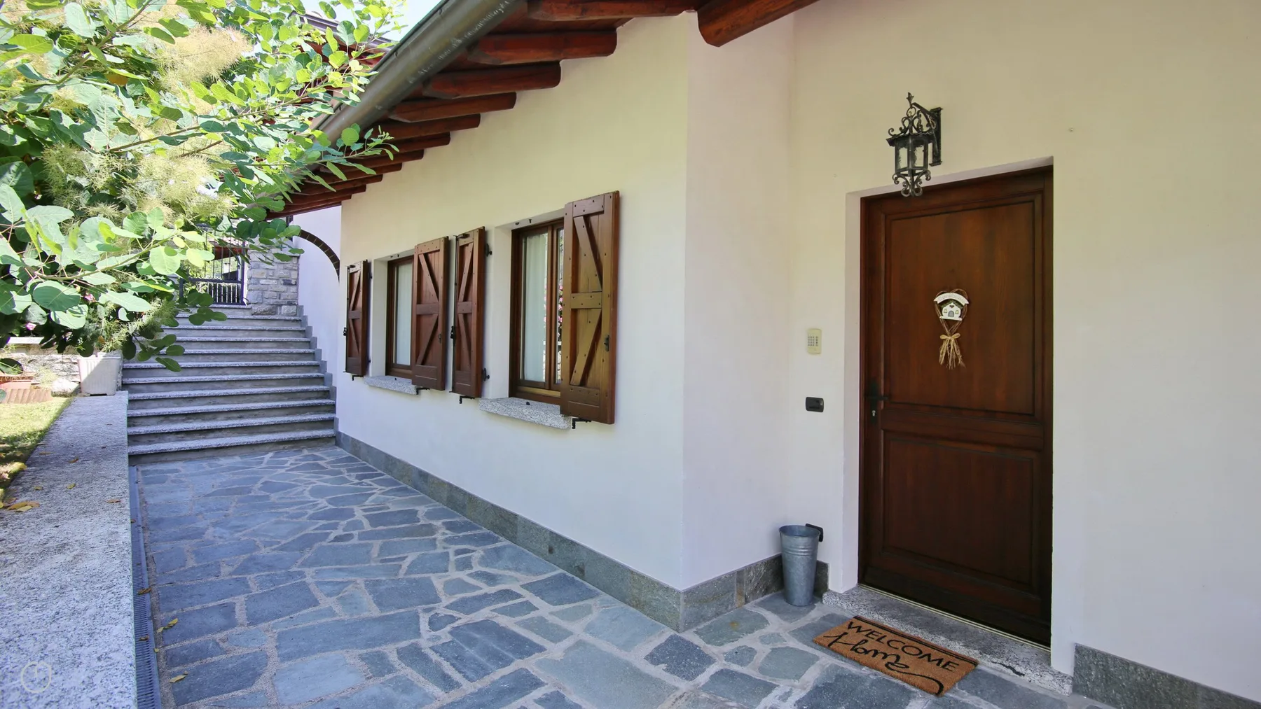 A stone-paved walkway runs beside a white building with wooden shutters and a door.