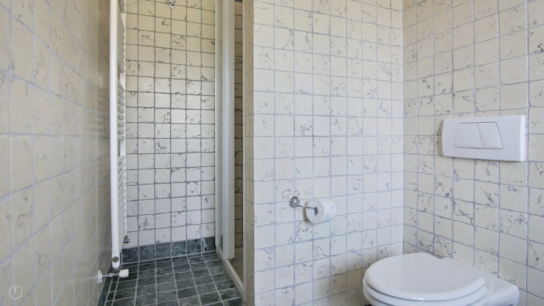 A tiled bathroom with a toilet beside a narrow shower area.