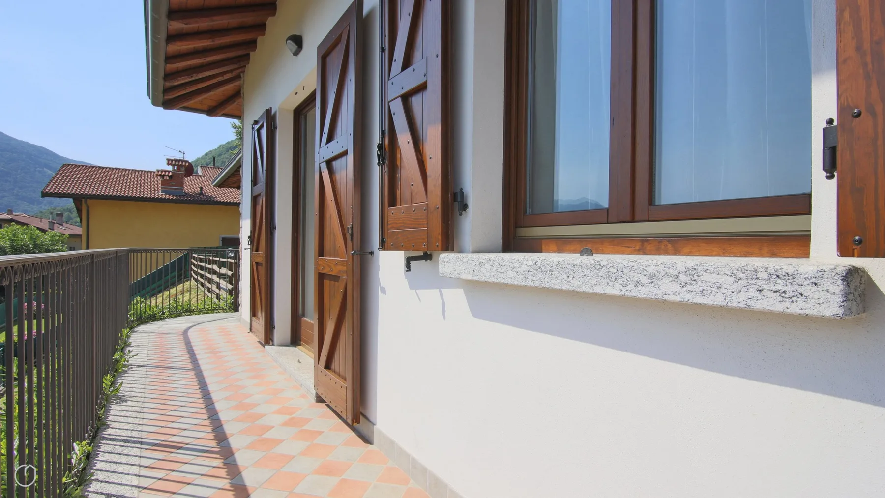 A narrow exterior walkway runs beside a building with wooden shutters and a railing.