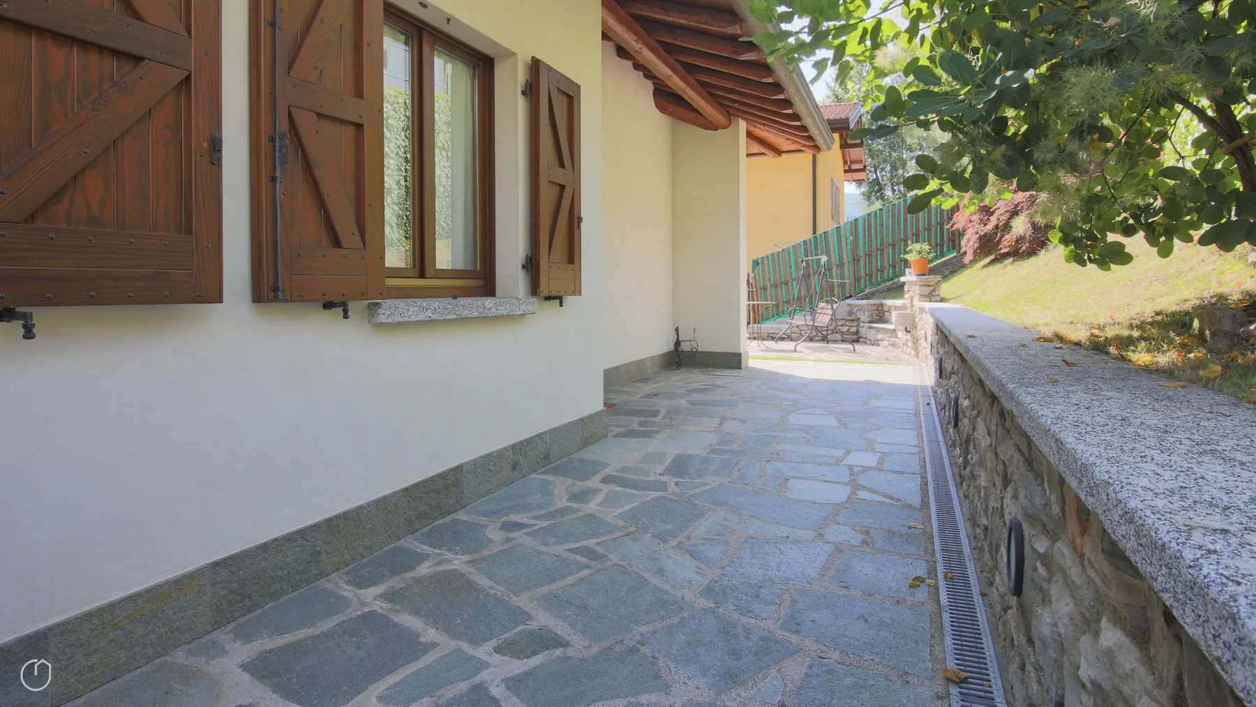 A stone-paved outdoor walkway runs alongside a light-colored building with wooden shutters.