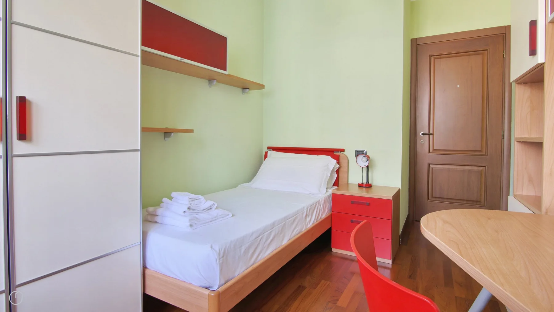 A compact bedroom with a single bed, red bedside table, and wooden door.