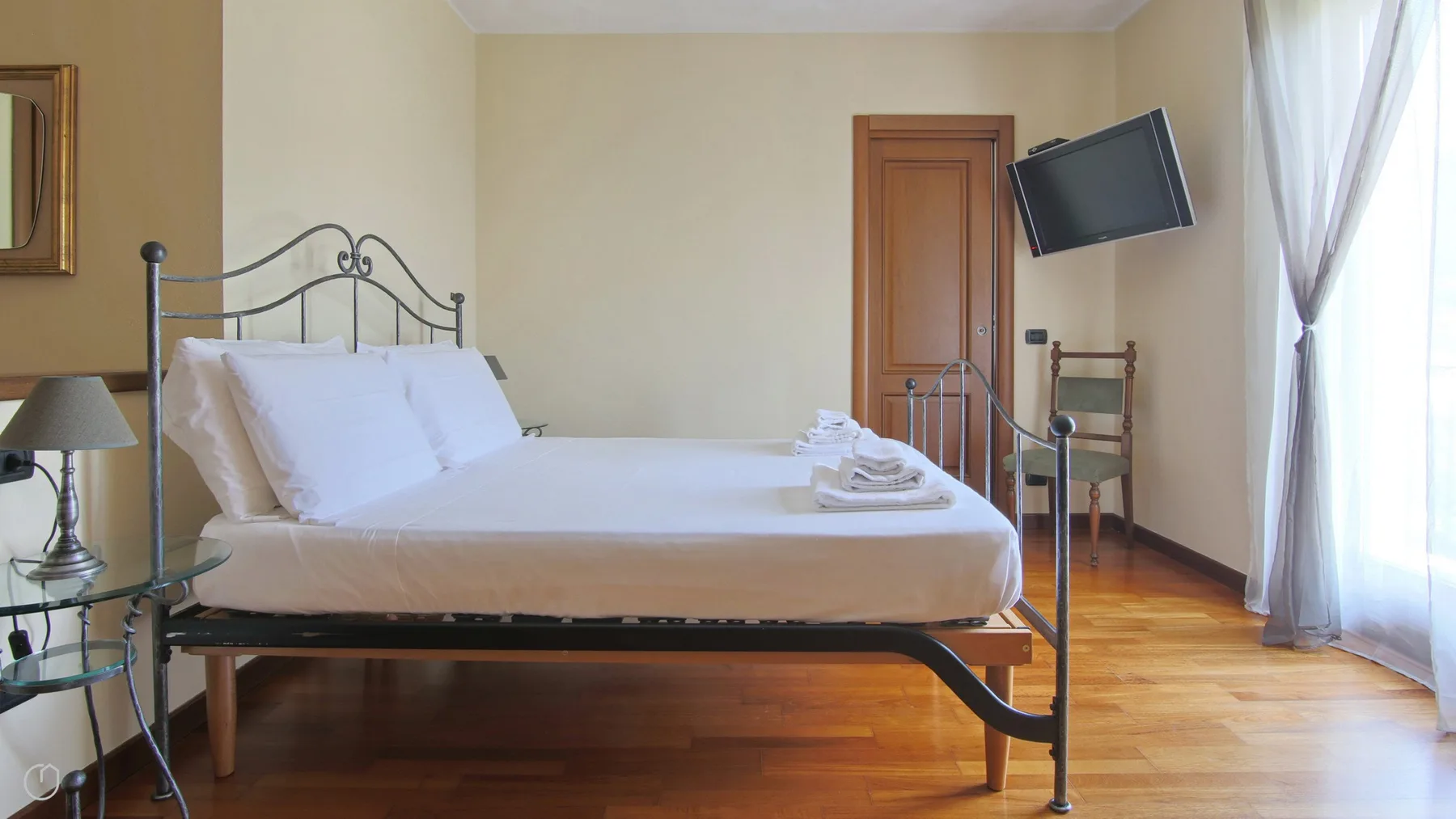 A bedroom with a neatly made bed and a television mounted near the window.