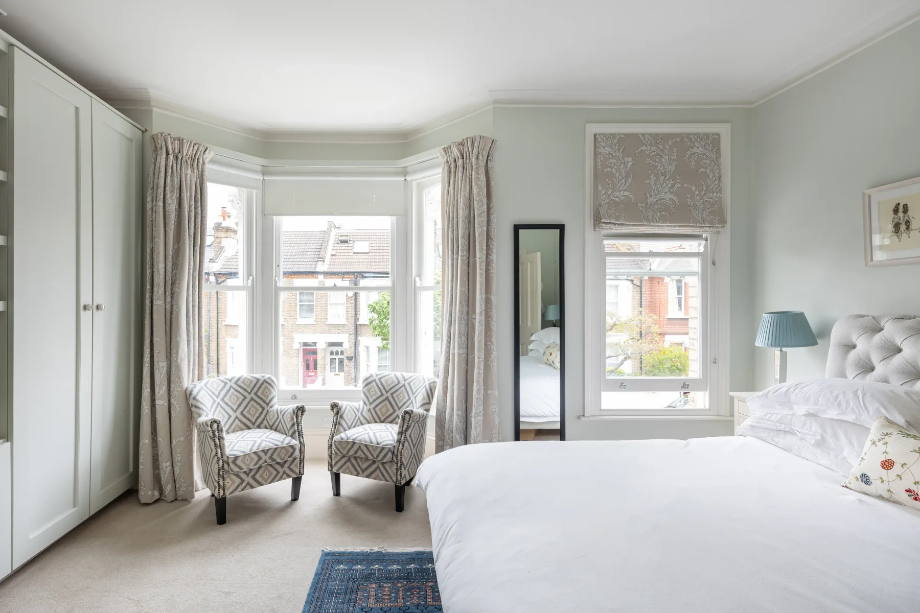 A bright bedroom with a large bed and two patterned armchairs by the windows.