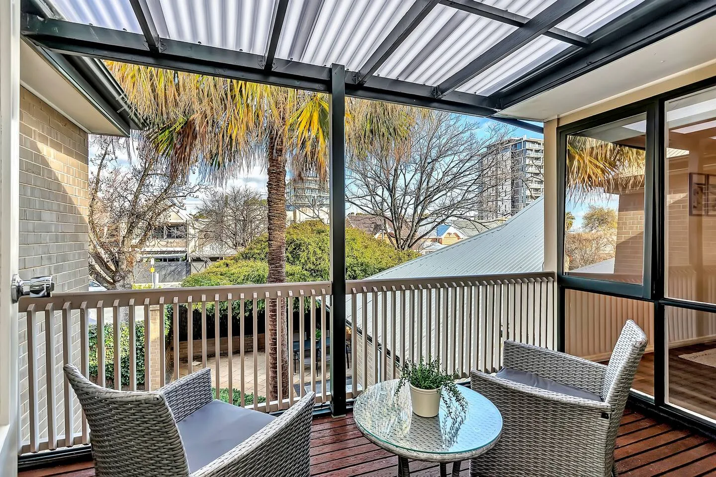 A covered balcony has seating around a small glass table, with trees visible beyond the railing.
