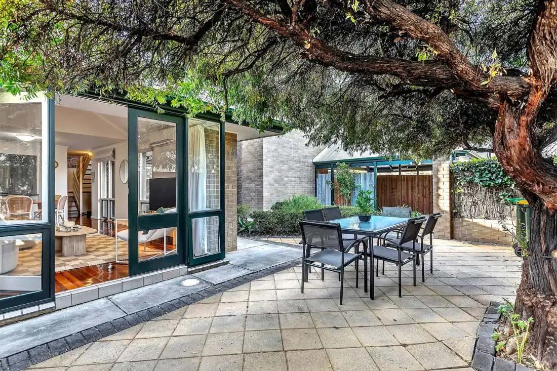 A paved outdoor dining area with a table and seating beneath a large tree.