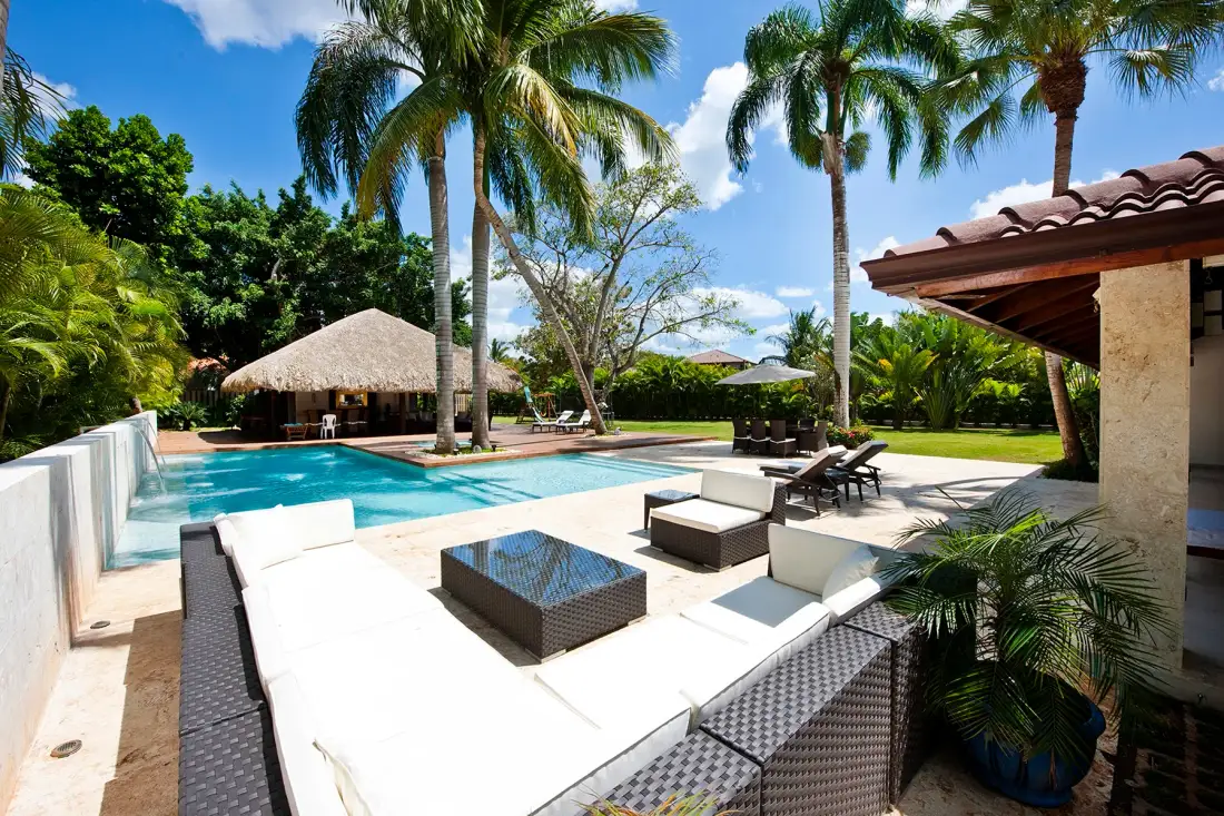 Un patio junto a la piscina con asientos y palmeras en un jardín tropical.