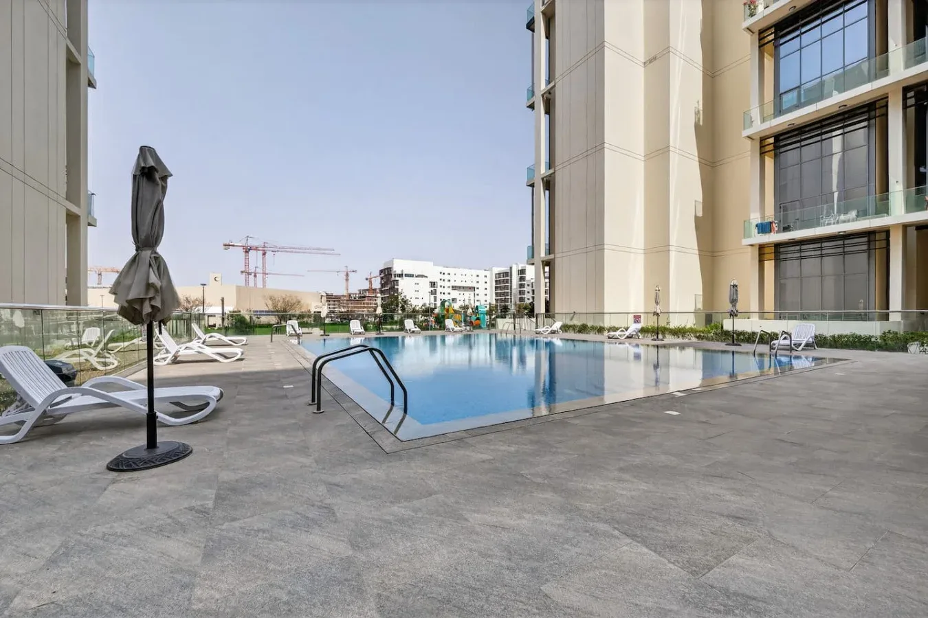 An outdoor swimming pool sits between modern buildings, with lounge chairs on the surrounding deck.