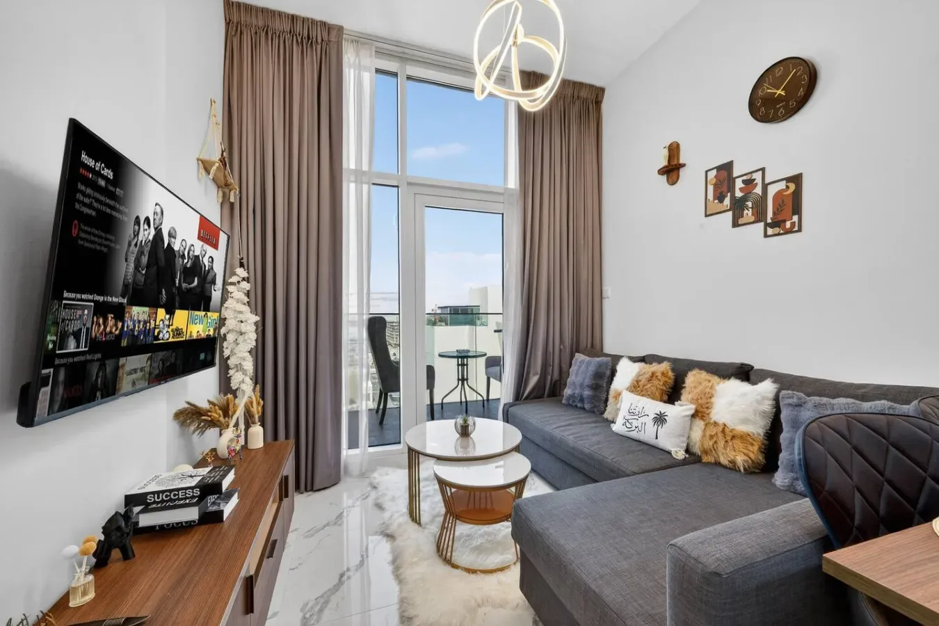 A bright living room with gray seating, a wall-mounted television, and tall windows.