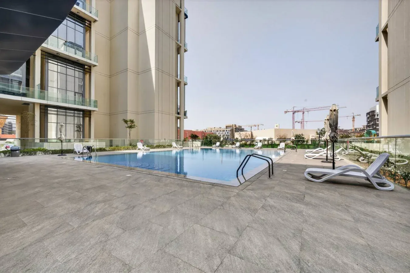 An outdoor swimming pool sits beside modern buildings, with loungers along the deck.