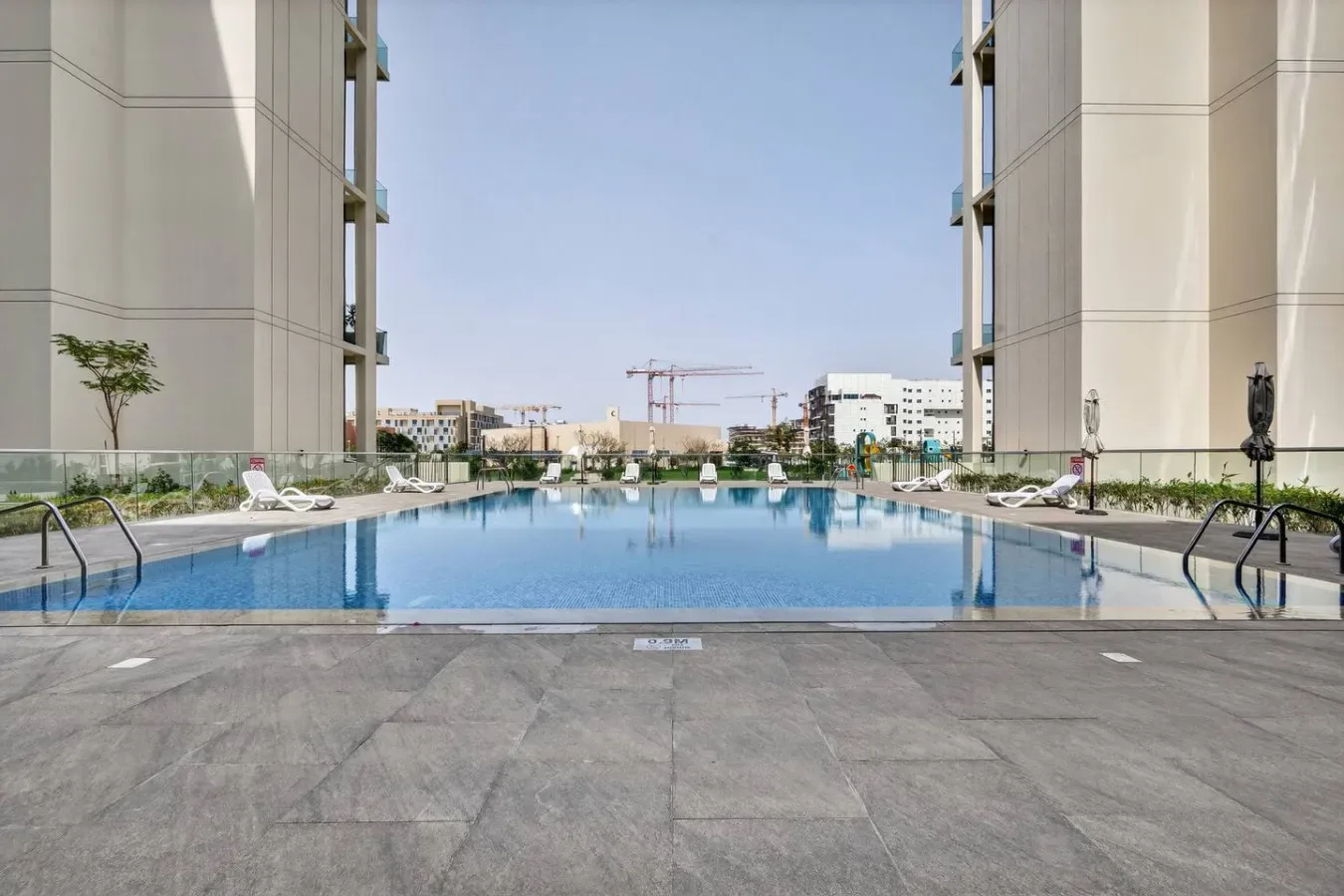 An outdoor swimming pool sits between tall buildings, with paved space along the edge.