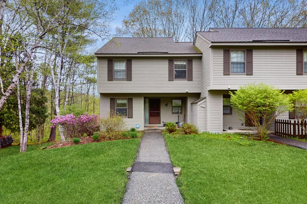 A two-story house stands behind a paved walkway, with green lawns and trees.
