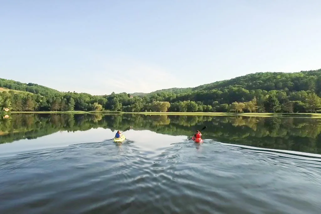 Two kayakers paddle across a calm lake beneath wooded hills.