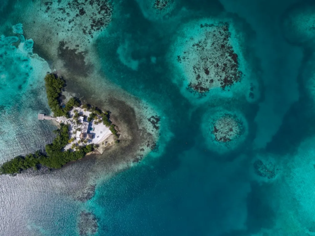 An aerial view shows a small tropical island surrounded by turquoise water and coral reefs.