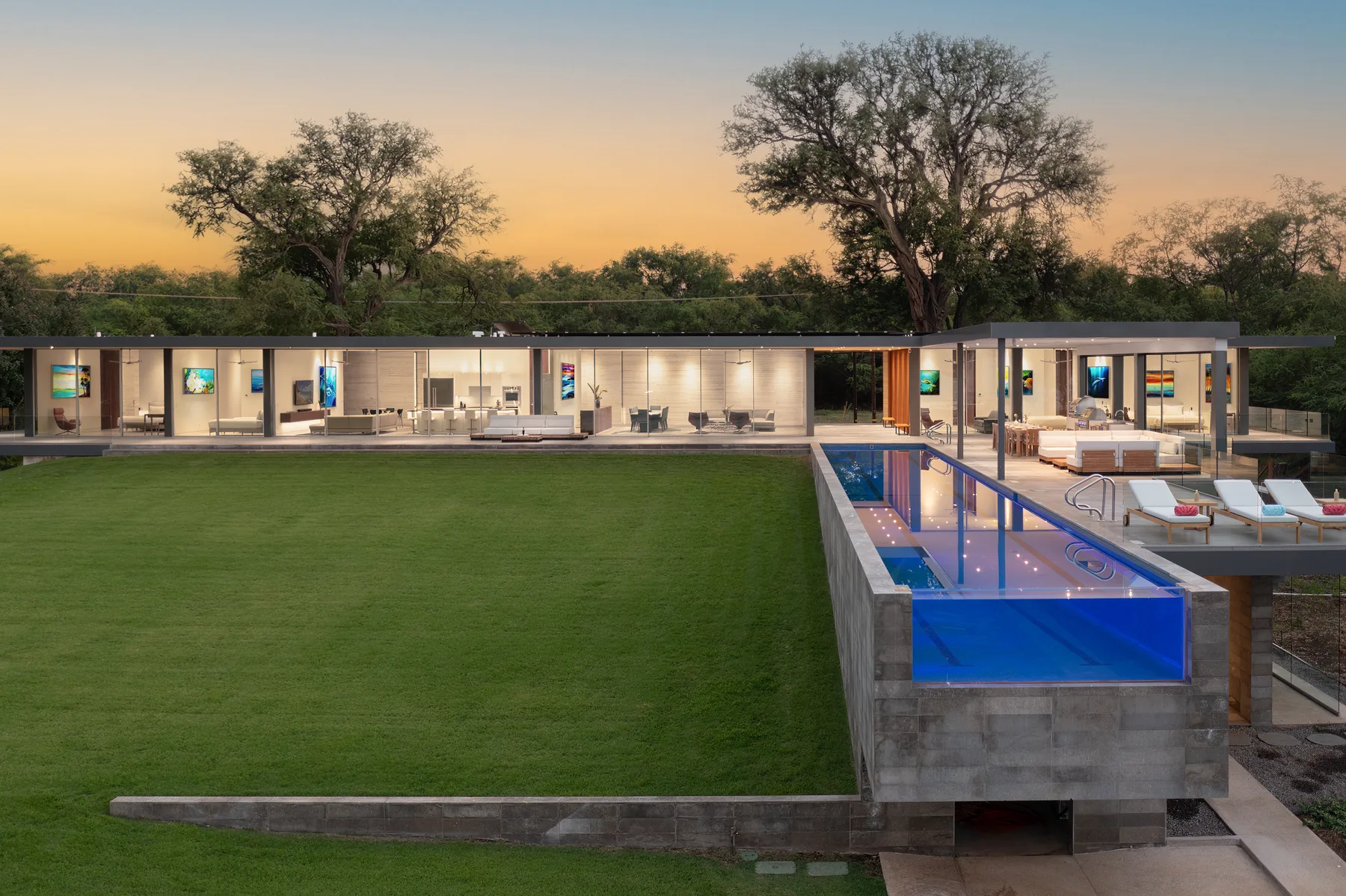A modern house faces a broad lawn, with a blue-lit pool in the foreground.