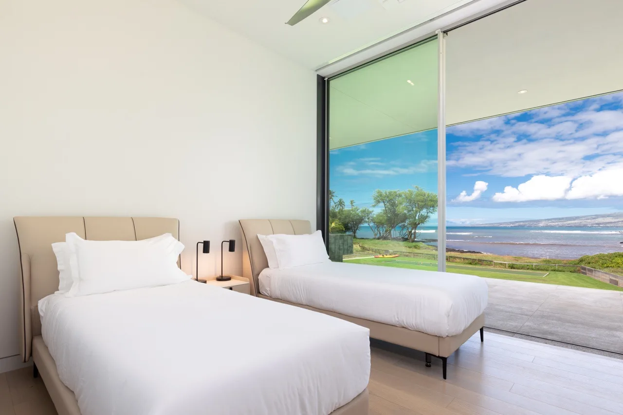 A bright bedroom with two beds beside a large window overlooking the water.
