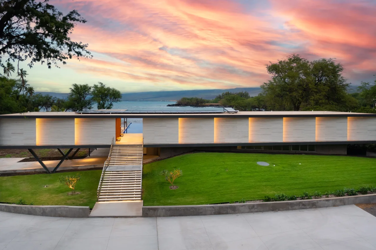 Un edificio moderno da a un césped verde y al agua bajo un cielo colorido al atardecer.