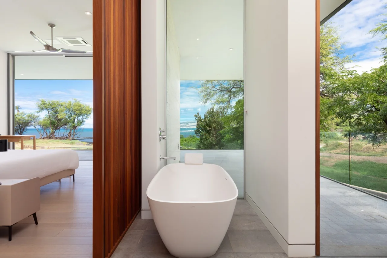 A freestanding bathtub sits beside a glass door overlooking greenery and the sea.