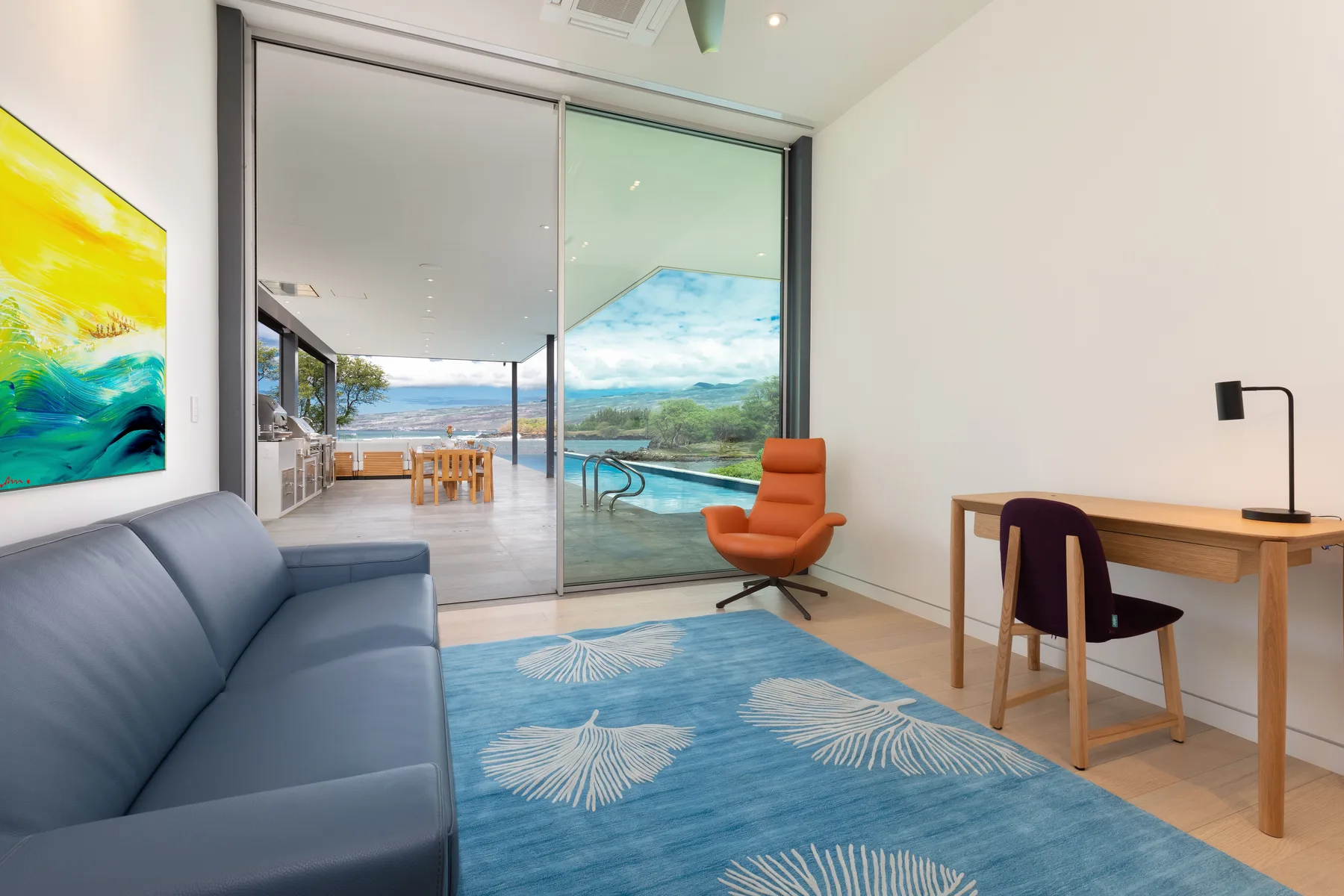 A bright living area with blue seating, a desk, and large glass doors overlooking a pool.